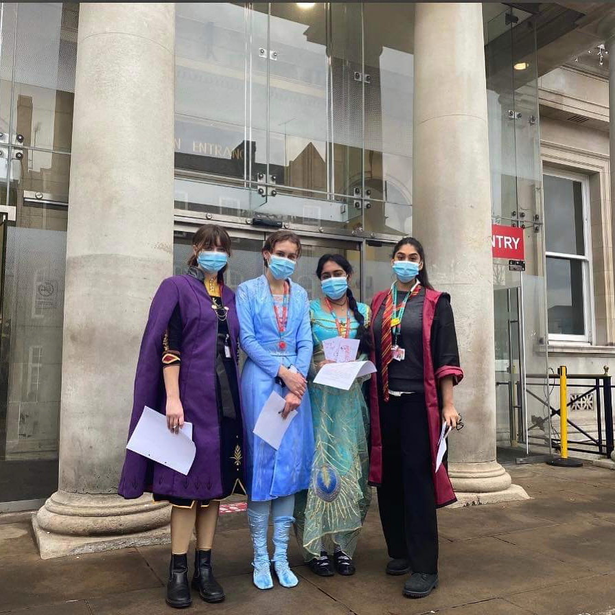 Four young women standing in front of a hospital in character costumes - a wizard, two princesses and a Harry Potter character.